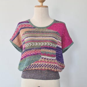May include: A vibrant, patchwork-style knitted top with short sleeves. The top showcases a variety of patterns and colours, including pink, green, purple, and beige. It has a round neckline and a small pocket. The bottom hem features a checkered pattern.