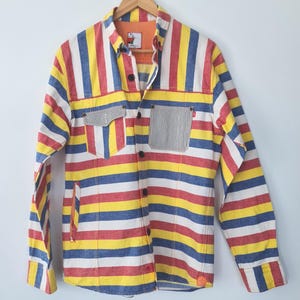 May include: A long-sleeved shirt with vertical stripes in red, yellow, blue, and white. The shirt has two front pockets, one with a striped pattern and the other with a solid color. The shirt is button-down and has a collar.