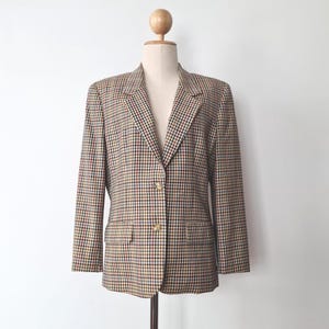 May include: A checkered blazer in a classic style, displayed on a mannequin. The blazer features a pattern of small squares in brown, red, black, and yellow. It has two front pockets and gold-colored buttons.