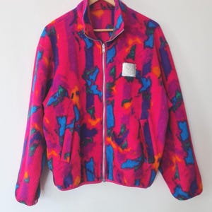 May include: A vibrant, multi-colored fleece jacket with a full zipper closure. The jacket features a bold pattern of pink, blue, purple, and orange. A small silver square is on the chest. The jacket is hanging on a wooden hanger.