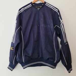 May include: Navy blue baseball jacket with white stripes and the word "MARIN" printed on the chest. The jacket has a zipper closure and a Mizuno logo on the sleeve. The jacket is hanging on a wooden hanger.
