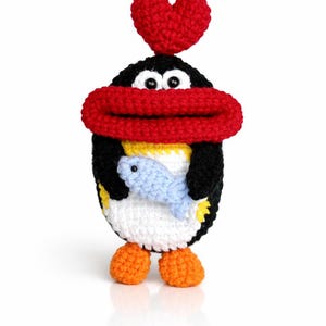 May include: A handmade crochet penguin with a red heart on its head and a red scarf. The penguin is holding a blue fish. It has orange feet and a white belly. The penguin is made of black, white, yellow, and orange yarn.