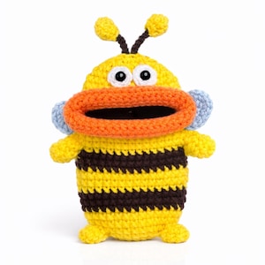 May include: A handmade crochet bee toy in yellow and brown stripes. The bee has a large orange mouth, blue cheeks, and black and white eyes. The bee has small yellow antennae and is standing upright.