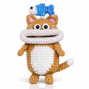 May include: A handmade crochet cat toy in shades of brown and white, with a blue and white fish on its head. The cat has large, round eyes and a wide, white mouth. The toy is standing upright on a white surface.