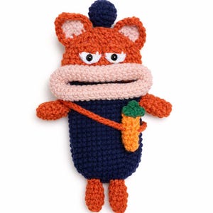 May include: A handmade crochet creature in orange, navy blue, and pink. The creature has a large mouth, white eyes, and a small carrot in its hand. The creature is wearing a navy blue outfit and has orange feet.