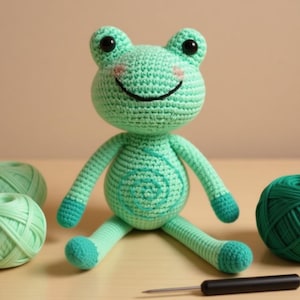 May include: A handmade crochet frog doll in shades of green, with a smiling face and rosy cheeks. The frog has a spiral design on its belly and is sitting on a wooden surface. Yarn balls and a crochet hook are also visible.