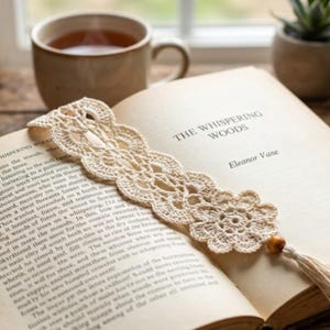 Può includere: Un segnalibro all'uncinetto color crema con un intricato motivo in pizzo e un fiore all'estremità. Il segnalibro è decorato con una piccola perla di legno e una nappa abbinata, appoggiato sulle pagine aperte del libro The Whispering Woods.