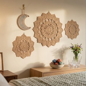 May include: A collection of beige crochet wall hangings in various sizes and shapes, including a crescent moon and circular doily-like designs. The decor is displayed on a light-colored wall, creating a bohemian aesthetic. A wooden nightstand with a bowl of yarn and a vase of flowers is in the foreground.