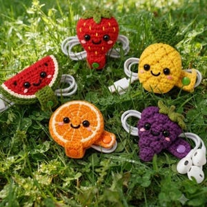May include: Five crocheted fruit-shaped cable organizers featuring a red strawberry, a yellow pineapple, a purple grape cluster, an orange slice, and a watermelon wedge. Each piece has a smiling face, black eyes, and a snap button to secure white cords.