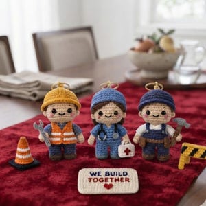 May include: Three crocheted dolls representing different professions, including a construction worker with a yellow hard hat, a medical professional with a stethoscope, and a laborer with a hammer, alongside a sign reading WE BUILD TOGETHER.