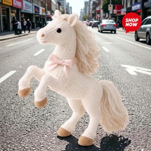 May include: A handmade, off-white crochet horse with a light pink bow around its neck. The horse has a flowing mane and tail, and brown hooves. The horse is posed rearing up on its hind legs.
