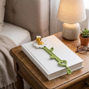 May include: A handmade crochet bookmark featuring a white daisy flower with a yellow center, topped with a small yellow and black striped bee. The stem is green with three leaves and a heart-shaped base, resting on a closed white hardcover book.