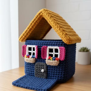 Könnte beinhalten: Ein blaues Häuschen aus Häkelarbeit mit einem senfgelben Dach. Es hat rosa Fensterläden, weiße Fensterrahmen und Blumenkästen. Eine kleine, graue Tür mit einem weißen Knopf ist ebenfalls vorhanden. Handgefertigtes Deko-Haus.