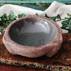 Może przedstawiać: Mała, okrągła ceramiczna miseczka o szorstkiej, teksturowanej brązowej powierzchni zewnętrznej i gładkim, błyszczącym ciemnoszarym wnętrzu. Misa posiada lekko nierówny, organiczny brzeg oraz widoczną sygnaturę twórcy na wewnętrznej ściance.