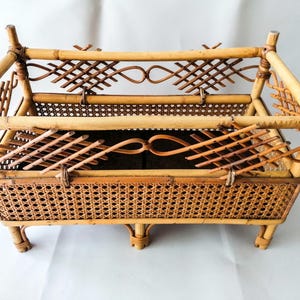 May include: A rectangular light brown bamboo and rattan magazine rack featuring a woven cane base and sides. The structure includes four vertical corner posts and decorative lattice detailing along the upper frame, supported by four small feet.
