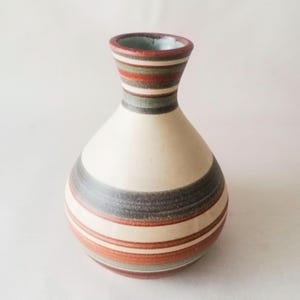 Puede incluir: Un jarrón de cerámica con cuerpo crema y rayas horizontales en tonos marrón, gris y rojo. El jarrón tiene una base redondeada y un cuello estrecho con un borde acampanado. El interior del borde es de color azul claro.