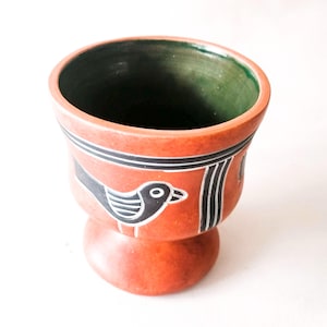 Könnte beinhalten: Eine terrakottafarbene Keramiktasse mit Sockel. Die Tasse hat ein schwarz-weißes Vogeldesign und schwarze Streifen. Der Innenraum ist grün. Die Tasse ist wahrscheinlich handgefertigt und hat eine rustikale Ästhetik.