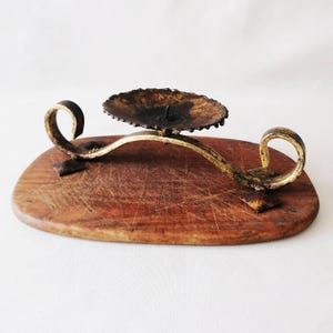 Puede incluir: Un candelabro de metal con un plato circular oscuro y texturizado, y un marco curvo de tono dorado con extremos en espiral. El soporte está montado sobre una base de madera plana y ovalada que muestra arañazos visibles y veta natural.