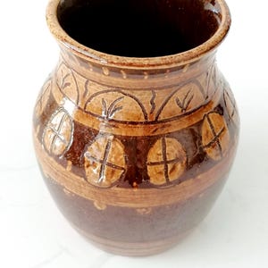 Puede incluir: Un jarrón de cerámica marrón hecho a mano con una abertura ancha. El jarrón presenta bandas decorativas con patrones de hojas y cruces en tonos marrones más claros. El interior es de color marrón oscuro.