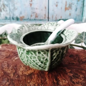 Puede incluir: Un azucarero de cerámica con forma de hoja de col verde, con un exterior texturizado y un interior blanco liso. Una pequeña cuchara de cerámica blanca con el mango de punta verde descansa dentro del cuenco, que tiene un borde festoneado.