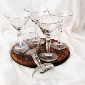 Puede incluir: Un juego de cinco copas de cóctel de cristal transparente con un delicado patrón floral grabado en el cuenco. Cada copa tiene un tallo fino y una base circular. Cuatro copas están de pie y una está colocada horizontalmente de lado.