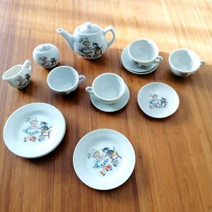 May include: A white ceramic tea set with a whimsical illustration of a girl and a black cat. The set includes a teapot, sugar bowl, creamer, cups, saucers, and plates. The set is arranged on a wooden surface.