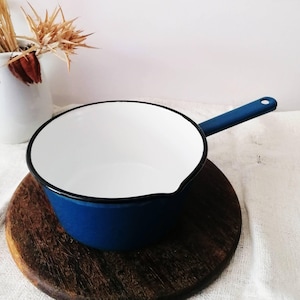 May include: A deep blue enamel saucepan with a white interior and a black rim. The pan features a long, straight blue handle extending from the side and a small pouring spout on the edge. The exterior finish is smooth and solid blue.
