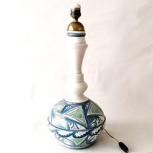 Può includere: Una lampada da tavolo in ceramica bianca con un portalampada e una base color ottone. La lampada ha una base bulbosa decorata con motivi geometrici blu e verdi e un collo bianco allungato.