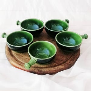 Puede incluir: Un conjunto de cinco cuencos redondos pequeños de cerámica verde, cada uno con un mango corto que se extiende desde el lateral. Los cuencos tienen un acabado brillante con un tono verde oscuro en el exterior y un interior verde claro, dispuestos en círculo.