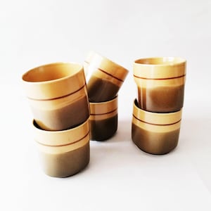 Puede incluir: Seis tazas de cerámica apiladas. Las tazas son cilíndricas con un diseño de dos tonos. La parte superior es de color beige claro, el medio es marrón oscuro y una fina raya marrón separa los dos colores.
