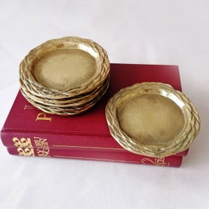 Puede incluir: Una pila de cuatro posavasos circulares de latón y un posavasos individual, cada uno con un borde texturizado en forma de cuerda. Los posavasos descansan sobre un libro de tapa dura rojo con letras doradas visibles en el lomo del libro.
