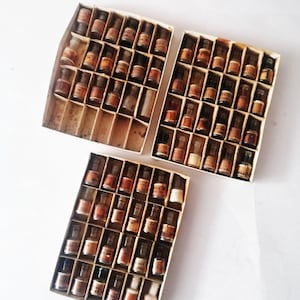Op de afbeelding: Drie open kartonnen dozen gevuld met kleine glazen flesjes. De flesjes zijn gerangschikt in rijen en kolommen, elk met een etiket. De flesjes hebben bruine en amberkleurige vloeistof. De dozen staan op een wit oppervlak.