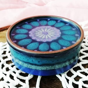 May include: A round ceramic lidded container featuring a deep blue and turquoise glaze. The lid displays a central light purple floral pattern surrounded by a ring of teal petal shapes. The base has horizontal bands of blue and speckled light blue.