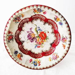 Puede incluir: Una bandeja de servicio redonda de metal con un centro color crema y un vibrante ramo de flores rojas, moradas y amarillas. La bandeja tiene un borde rojo festoneado decorado con filigrana dorada intrincada y pequeños detalles florales.