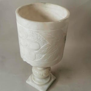 Puede incluir: Un cáliz de mármol blanco con diseños florales tallados. La copa tiene una abertura ancha y se asienta sobre una base de pedestal. El cáliz mide aproximadamente 18 cm de altura.