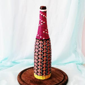 Op de afbeelding: Een decoratieve glazen fles omwikkeld met kleurrijk geweven draad. De hals heeft horizontale strepen in felroze, zwart en wit. Het lichaam heeft een zwart rasterpatroon over horizontale banden in rood, geel, blauw en groen. De basis is geel.