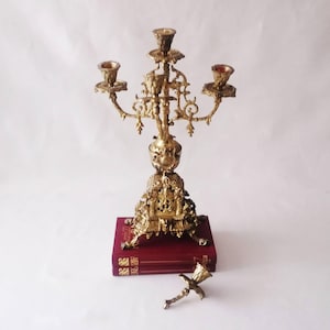 May include: A gold-toned metal candelabra with four candle holders, featuring intricate scrollwork and ornate detailing on the base and arms. The candelabra rests on two stacked dark red books, with a single detached candle holder piece nearby.