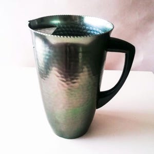 Puede incluir: Una jarra plateada con textura martillada y asa negra. La jarra tiene boca ancha y asa curva, adecuada para servir bebidas. El diseño general es sencillo y elegante.