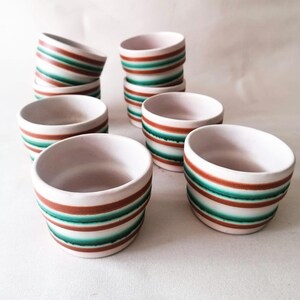 Puede incluir: Un conjunto de ocho cuencos pequeños de cerámica blanca. Cada cuenco está adornado con rayas horizontales en tonos marrón y verde azulado. Los cuencos están dispuestos en pilas e individualmente, mostrando su diseño.