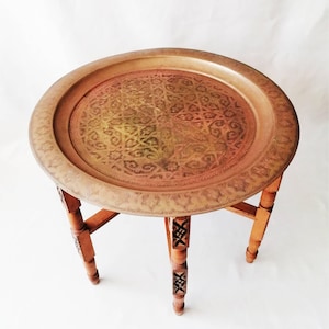 Op de afbeelding: Een ronde tafel met een cirkelvormig metalen dienblad als tafelblad, voorzien van een ingewikkeld gegraveerd geometrisch patroon in een messingkleur. De tafel rust op vier houten poten, elk met uitgesneden details langs de verticale lengte.