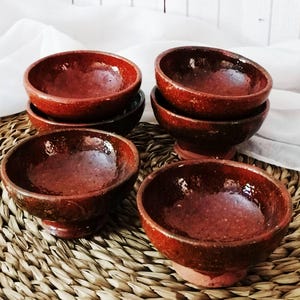 Op de afbeelding: Een set van vier kleine, ronde, diepe kommen met een glanzend, donker roodbruin gespikkeld glazuur. Elke kom heeft een brede opening en een korte, stevige voet, met een glad, reflecterend binnenoppervlak en een textuur aan de buitenkant.