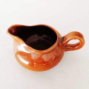 Puede incluir: Una pequeña jarra de cerámica de forma redonda con un esmalte brillante de color ámbar cálido. El recipiente presenta un pico vertedor ancho y curvado en un lado y un asa robusta en forma de bucle en el lado opuesto, con acabado liso.