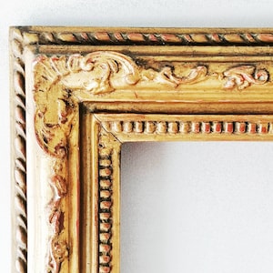 Op de afbeelding: Een rechthoekige fotolijst met een gouden afwerking en zichtbare rode ondertonen. De lijst heeft ingewikkeld houtsnijwerk in de hoeken, een touwachtige buitenrand en een kralenranddetail langs de binnenkant van de opening.
