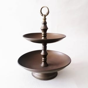 Könnte beinhalten: Bronze-farbener, zweistöckiger Servierständer mit dekorativem Griff. Der Ständer hat einen runden Sockel und zwei runde Tabletts, ideal zum Präsentieren von Speisen.