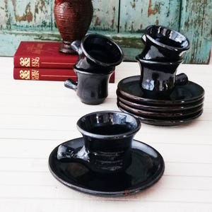 Puede incluir: Un juego de tazas de cerámica negra brillante con sus platos a juego. Las tazas tienen una pequeña asa redondeada en el lateral. Algunas tazas están apiladas, mientras que una se encuentra colocada individualmente sobre un plato.
