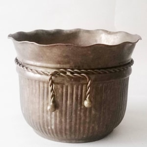 Puede incluir: Una maceta marrón metálica de estilo vintage con un borde festoneado y detalles acanalados verticales. La maceta presenta una banda en forma de cuerda alrededor del centro, con detalles decorativos de cuerda y bola. Esta maceta decorativa es adecuada para uso en interiores o exteriores.