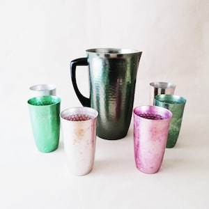 May include: A large dark green hammered metal pitcher with a black handle, surrounded by six matching hammered metal tumblers in various colours including green, pink, and silver, all featuring a textured finish and cylindrical shape.