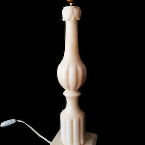 Puede incluir: Lámpara de mesa de mármol color crema con base estriada y centro bulboso. La lámpara tiene un cuello largo y esbelto y un casquillo de latón. Un cable blanco con un interruptor de encendido/apagado sale de la base.