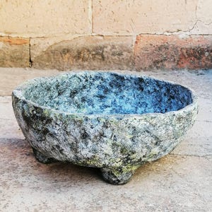 Puede incluir: Un cuenco redondo de perfil bajo con una superficie rugosa y texturizada en tonos gris moteado, negro y toques de verde. El cuenco cuenta con tres patas cortas y resistentes que sostienen su base y tiene una parte superior abierta y ancha.