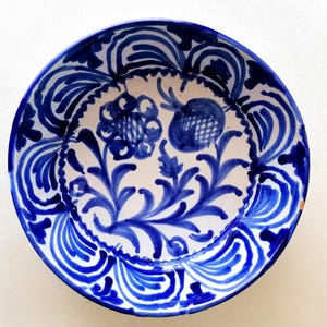 Op de afbeelding: Een ronde keramische kom met een witte basis en handgeschilderde kobaltblauwe bloem- en granaatappelmotieven. De binnenrand is versierd met herhalende blauwe gebogen patronen, terwijl het midden een gedetailleerd botanisch ontwerp toont.